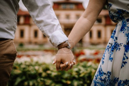Close-up of a couple holding hands, man in white shirt with watch, woman in floral dress, blurred building background.