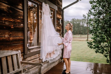 Valmiera, Latvia - August 10, 2023 - A bride in a pink robe admires her hanging wedding dress outside a wooden house.