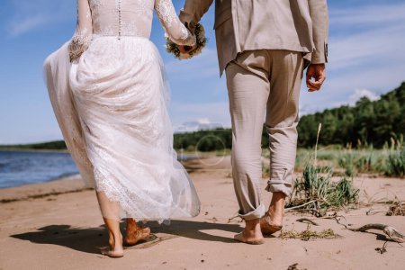 Valmiera, Latvia - August 10, 2023 - Barefoot bride and groom walking on beach, bride holding up dress, groom's trouser rolled up, both holding hands.