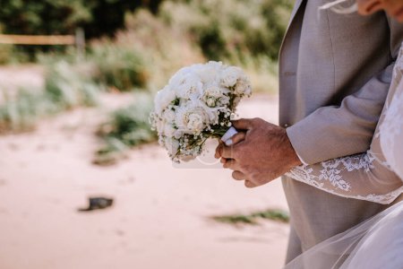 Photo for Valmiera, Latvia - August 10, 2023 - Close-up of a groom holding a bridal bouquet of white roses at a beach wedding. - Royalty Free Image