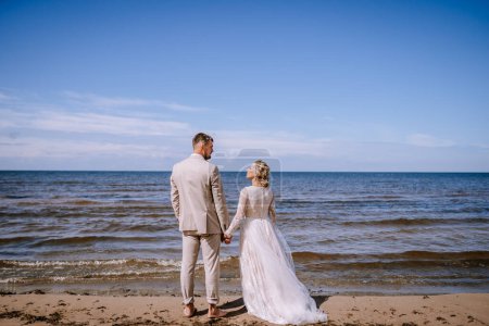 Foto de Valmiera, Letonia - 10 de agosto de 2023 - Novia y novio tomados de la mano en la playa mirando al mar - Imagen libre de derechos