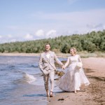 Valmiera, Latvia - August 10, 2023 - Bride and groom walking hand in hand along a sandy beach, with waves and trees in the background.