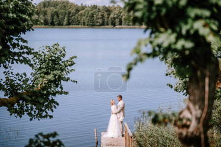 Foto de Valmiera, Letonia - 10 de agosto de 2023 - Una pareja en traje de novia se abraza en un muelle de madera junto al lago, enmarcado por árboles. - Imagen libre de derechos
