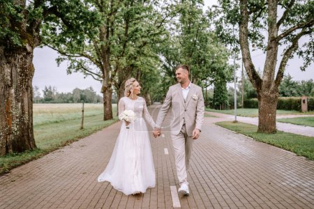 Photo for Valmiera, Latvia - August 10, 2023 - A bride and groom walking hand in hand on a cobblestone path lined with tall trees, exchanging smiles. - Royalty Free Image