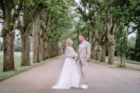 Valmiera, Latvia - August 10, 2023 - A bride and groom stand on a tree-lined path, looking at each other, dressed in wedding attire.