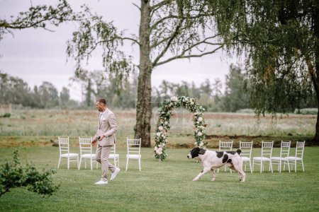 Photo for Valmiera, Latvia - August 10, 2023 - A groom walking beside a floral arch at a garden wedding with a dog passing by. - Royalty Free Image