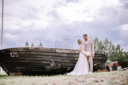 Valmiera, Latvia - August 10, 2023 - Bride and groom holding hands in front of an old wooden boat, with a cloudy sky above.