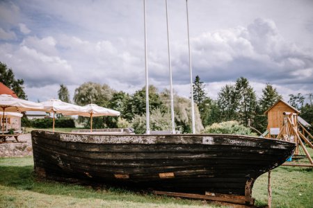 Valmiera, Lettland - 10. August 2023 - Altes Holzboot auf Gras mit Sonnenschirmen und einem Spielset im Hintergrund unter bewölktem Himmel.