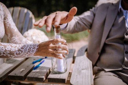 Valmiera, Latvia - August 10, 2023 - Close-up of a bride and groom's hands placing a note into a bottle on a wooden table.