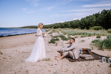 Foto de Valmiera, Letonia - 10 de agosto de 2023 - Novia y novio en una playa, con el novio sentado y la novia de pie, ambos sosteniendo copas de vino. - Imagen libre de derechos
