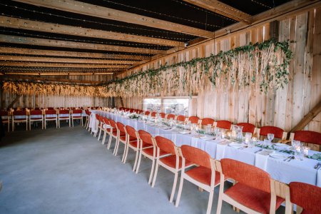 Photo for Valmiera, Latvia - August 10, 2023 - Rustic wedding banquet hall setup with a long table, red chairs, hanging lights, and floral decorations. - Royalty Free Image