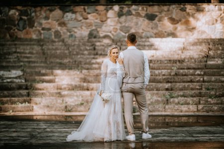 Valmiera, Lettland - 10. August 2023 - Braut blickt Kamera über die Schulter des Bräutigams auf verwitterter Holzbühne, Steinkulisse.