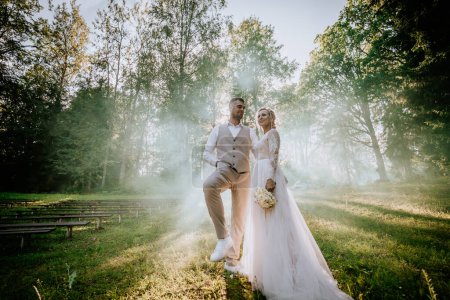 Photo for Valmiera, Latvia - August 10, 2023 - Bride and groom stand amid misty forest clearing, sunbeams filtering through trees. - Royalty Free Image
