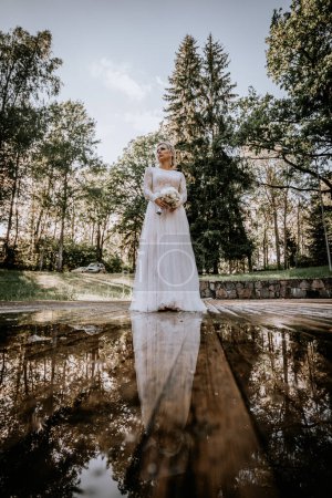 Valmiera, Lettland - 10. August 2023 - Eine besinnliche Braut steht auf einer spiegelnden Oberfläche mit Bäumen im Hintergrund, ihr Bild spiegelt sich im Wasser unten.