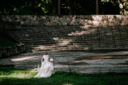 Valmiera, Letonia - 10 de agosto de 2023 - Una novia con un vestido blanco sostiene un ramo, de pie sola en un gran escenario de madera con escalones de piedra en el fondo.