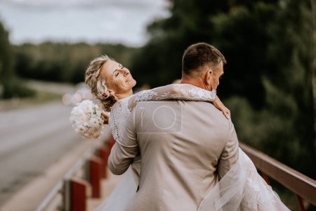 Valmiera, Letonia - 10 de agosto de 2023 - La novia se inclina de nuevo en los brazos del novio, ambos mirando felices, junto a una barandilla, árboles en el fondo.