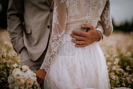 Foto de Valmiera, Letonia - 10 de agosto de 2023 - Acercamiento de la mano de un novio en la cintura de una novia, el detalle de su vestido de encaje y su ramo de bodas. - Imagen libre de derechos