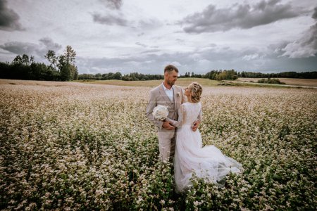 Valmiera, Lettonie - 10 août 2023 - Couple de mariés debout dans un champ de fleurs blanches sous un ciel dramatique.