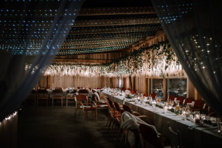 Valmiera, Letonia - 10 de agosto de 2023 - Elegante salón de banquetes con largas mesas de comedor, decorado con flores y luces de hadas, listo para un evento festivo.