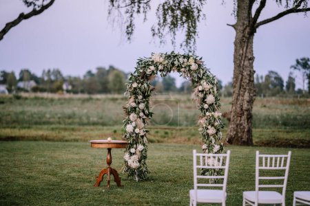 Valmiera, Lettland - 10. August 2023 - Hochzeitskulisse im Freien mit dekorativem Blumenbogen, Vintage-Tisch und weißen Stühlen auf dem Rasen, Bäume im Hintergrund.