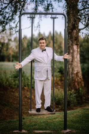 Valmiera, Letonia - 10 de agosto de 2023 - Hombre de traje blanco de pie con los brazos extendidos en un marco de metal al aire libre, mirando divertido.