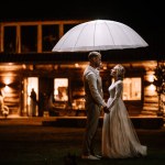 Valmiera, Latvia - August 10, 2023 - Bride and groom holding hands under an umbrella at night, with a cabin in the background.
