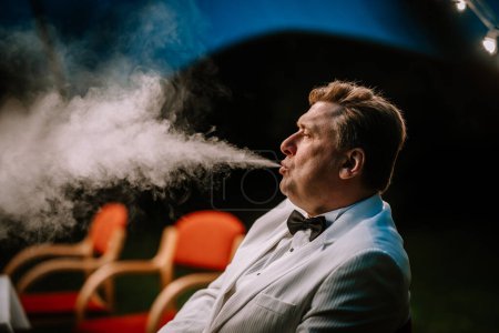 Valmiera, Lettland - 10. August 2023 - Mann im Smoking atmet draußen Rauch aus, im Hintergrund Stühle.