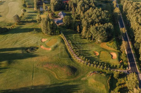 Foto de Valmiera, Letonia - 12 de agosto de 2023 - Aerial shot of golf course with sand bunkers, pond, trees, and adjacent road at sunset. - Imagen libre de derechos