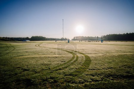 Téléchargez les photos : Valmiera, Lettonie - 12 août 2023 - Terrain de golf couvert de rosée au lever du soleil avec traces de pneus et arbres lointains sous un ciel dégagé. - en image libre de droit