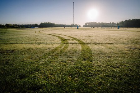 Téléchargez les photos : Valmiera, Lettonie - 12 août 2023 - Terrain de golf couvert de rosée au lever du soleil avec traces de pneus et arbres lointains sous un ciel dégagé. - en image libre de droit