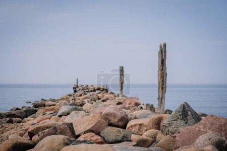 Tuja, Latvia - August 14, 2023 - Rocky jetty with weathered wooden posts leading into calm sea, clear sky.