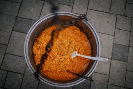 Dobele, Latvia - Augist 18, 2023 - Top-down view of grated carrots in a large metal pot with a plastic stirrer, hanging by a chain over pavement.