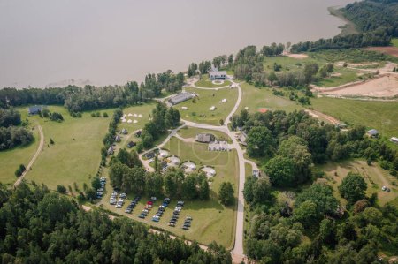 Dobele, Lettland - 18. August 2023 - Luftaufnahme einer ländlichen Landschaft mit Gebäuden, Zelten, einem Parkplatz und einem Gewässer.