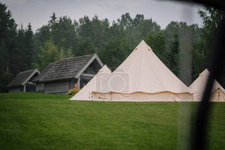 Dobele, Latvia - August 18, 2023 - A canvas bell tent on a rainy day, with pine trees and rustic cabins in the background.