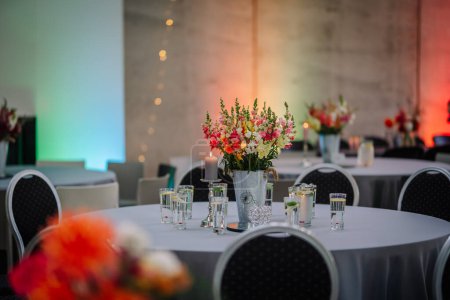 Dobele, Lettland - 18. August 2023 - Ein Blumengesteck auf einem Veranstaltungstisch mit Kerzen, Gläsern und weiteren Tischen in einem Raum mit farbiger Beleuchtung.