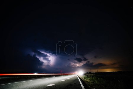 Dobele, Lettland - 18. August 2023 - Eine nächtliche Landschaft, die eine Straße mit Lichtstreifen eines fahrenden Fahrzeugs, einen stürmischen Himmel mit sichtbarem Blitz und einen beleuchteten Horizont zeigt..