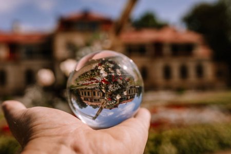 Valmiera, Latvia - August 19, 2023 - A crystal ball held in a hand shows an inverted, focused image of a historic building with lush greenery, set against a blurry background.