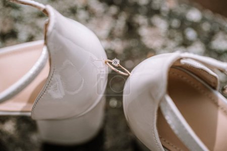 Valmiera, Latvia - August 19, 2023 - Close-up of elegant white wedding shoes with a diamond engagement ring placed between them, showcasing intricate detailing.