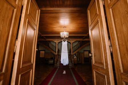 Valmiera, Lettland - 19. August 2023 - Ein Hochzeitskleid und Schuhe werden elegant in einem stattlichen Holzsaal mit zwei Treppen ausgestellt, hervorgehoben durch einen großen Kronleuchter.
