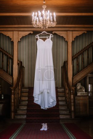 Valmiera, Latvia - August 19, 2023 - A bridal gown suspended from a chandelier in a luxurious wooden hallway with symmetrical staircases, enhancing its elegance.