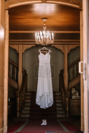 Valmiera, Latvia - August 19, 2023 - A wedding dress hangs from an ornate chandelier in a grand hallway with wooden walls and dual staircases, exuding an air of timeless elegance.