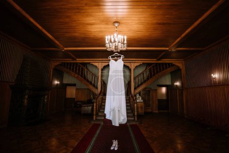 Valmiera, Latvia - August 19, 2023 - A wedding dress hangs majestically in a grand hall with a symmetrical wooden staircase, elegant chandelier, and opulent fireplace.