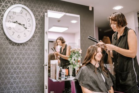 Valmiera, Latvia - August 19, 2023 - A hairstylist using a straightener on a smiling client's hair in a salon, with a clock and mirror in the background.
