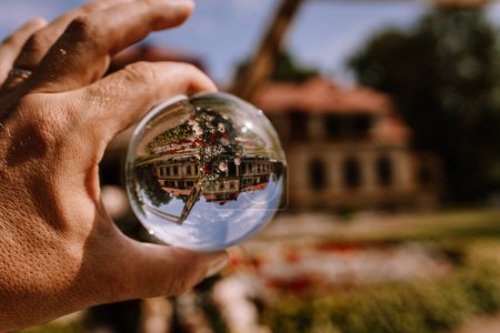 Valmiera, Letonia - 19 de agosto de 2023 - Una bola de cristal en una mano refleja vívidamente e invierte un edificio histórico, rodeado por un exuberante jardín y un cielo azul claro..