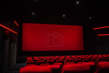Foto de Una sala de cine vacía con filas de asientos rojos frente a una gran pantalla roja, bañada en una luz roja malhumorada de la iluminación ambiental. - Imagen libre de derechos