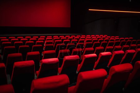 Filas de asientos rojos vacíos en una sala de cine oscura, iluminados por un brillo rojo de la pantalla, creando un ambiente malhumorado y atmosférico.