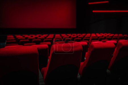 Dimly lit interior of a movie theater with rows of empty red seats facing a red screen, creating a moody, cinematic atmosphere.