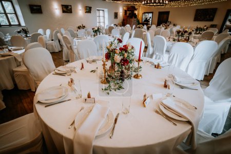 Valmiera, Latvia - August 19, 2023 - A wedding reception hall elegantly set with round tables covered in white linens, vibrant floral centerpieces, candles, and a string of fairy lights above.