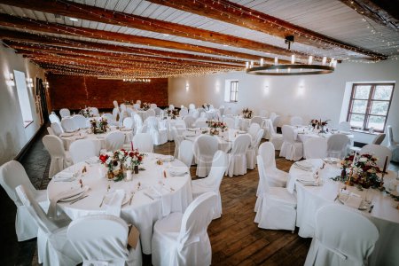 Valmiera, Lettland - 19. August 2023 - Indoor-Hochzeitslocation mit weiß drapierten Stühlen, runden Tischen mit floralen Herzstücken, Lichterketten an der Decke, rustikalem Ambiente.