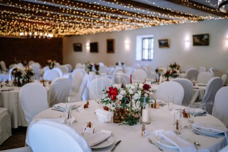 Valmiera, Letonia - 19 de agosto de 2023 - Elegantemente colocadas mesas de recepción de bodas con sillas blancas, manteles y vibrantes centros florales bajo luces centelleantes.